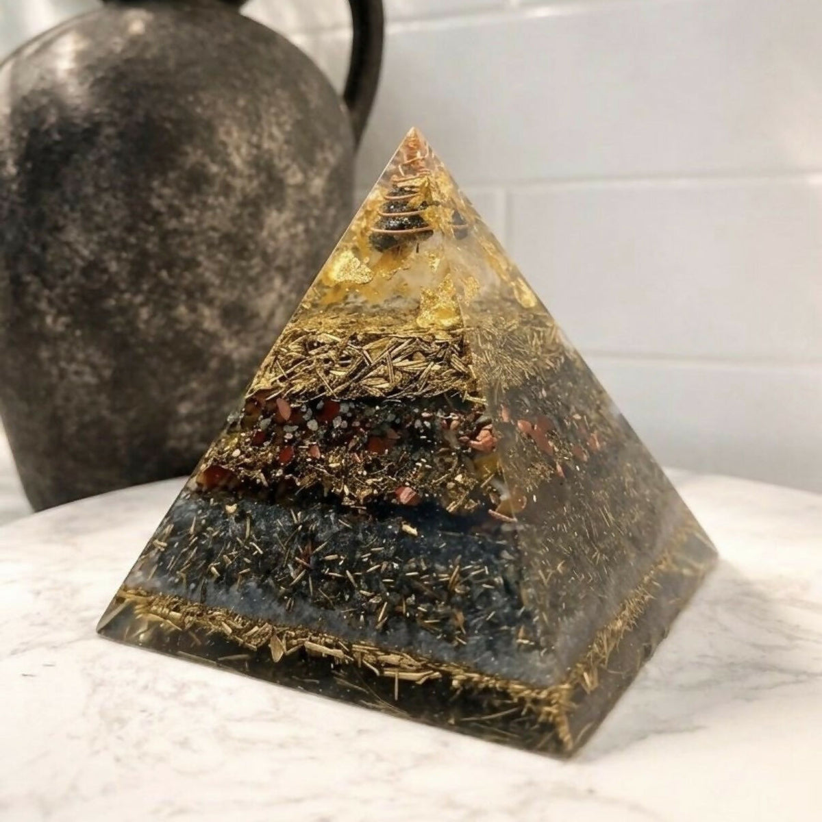 Pyramid on a white marble surface beside a dark ceramic jug, showing gold, red and grey layers