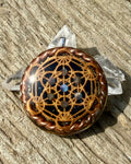 Metatron’s Cube Orgonite Pendant by Orgone Solutions – round dark pendant with a copper-coloured Metatron’s Cube design and small blue stones, edged with twisted copper wire, on weathered wood beside clear quartz points