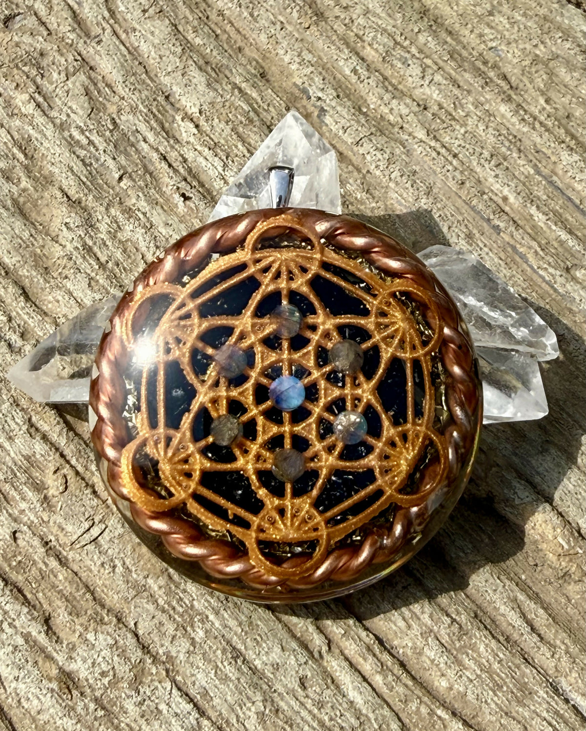 Metatron’s Cube Orgonite Pendant by Orgone Solutions – round dark pendant with a copper-coloured Metatron’s Cube design and small blue stones, edged with twisted copper wire, on weathered wood beside clear quartz points
