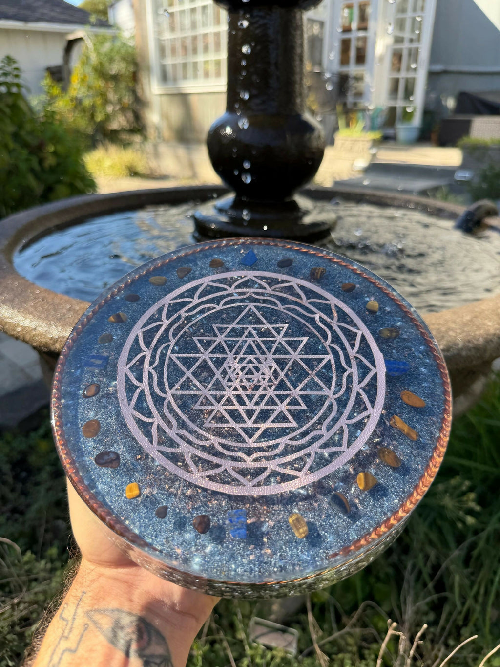 Top view of the plate held in front of a garden fountain, showing the Sri Yantra pattern framed by blue and amber stones