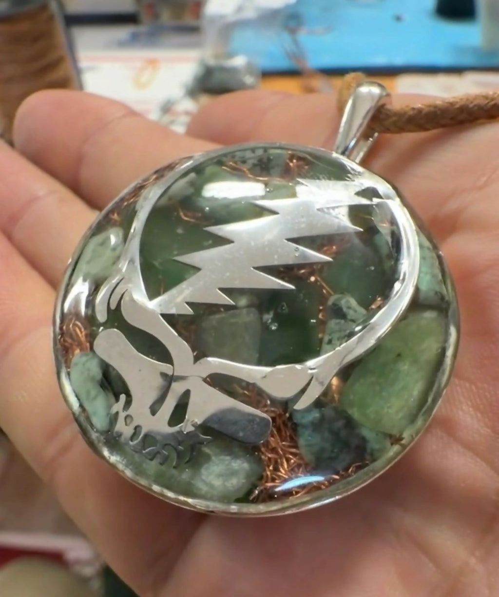 Stealie Jade & Peridot Pendant by Mark Ethan Designs – round domed pendant held in a hand, a silver skull-and-lightning-bolt design over green stones and copper, on a brown cord