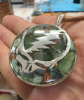 Stealie Jade & Peridot Pendant by Mark Ethan Designs – round domed pendant held in a hand, a silver skull-and-lightning-bolt design over green stones and copper, on a brown cord