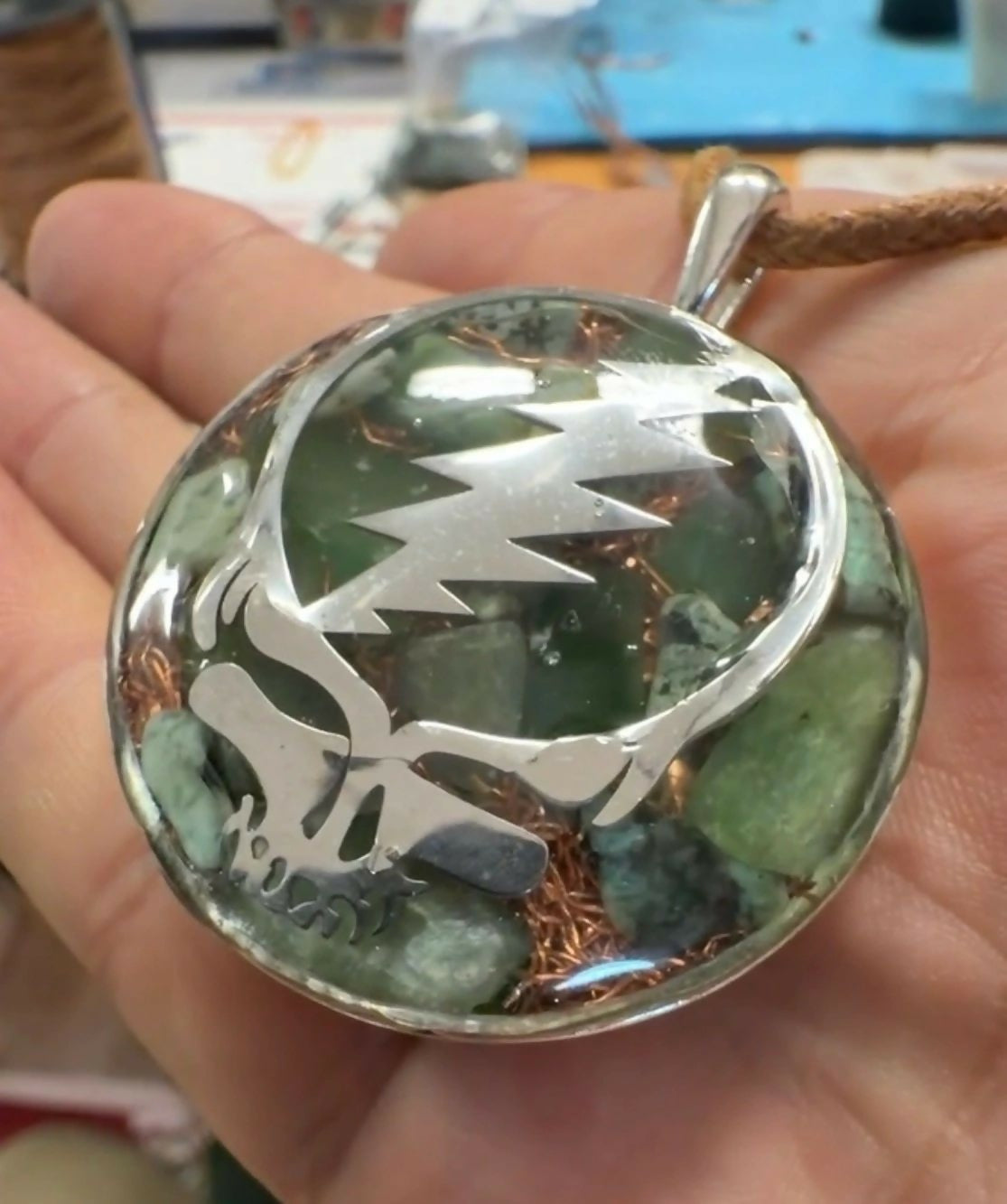 Stealie Jade & Peridot Pendant by Mark Ethan Designs – round domed pendant held in a hand, a silver skull-and-lightning-bolt design over green stones and copper, on a brown cord