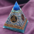 Small Tesla Orgonite Pyramid by Orgone Art Yvonne – pyramid with a blue sphere at the top over pale blue-green chips, gold triskele and spiral discs on dark red, copper band and turquoise base