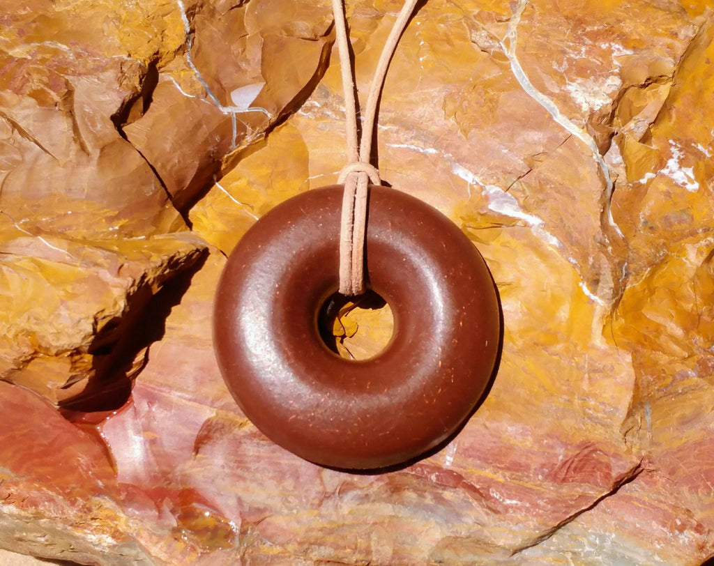Front view of the reddish-brown donut pendant hanging on its tan cord against layered red and yellow rock