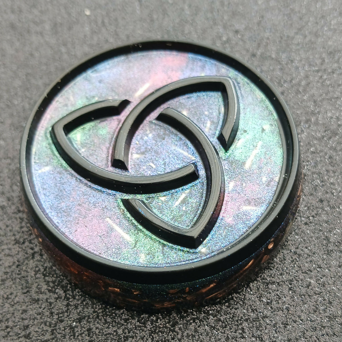 Close view of a round disc with a black raised triquetra knot over shimmering pastel blue and pink
