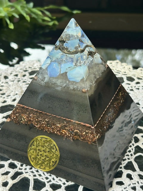 Chinese Dragon Orgonite Pyramid by Gaia's Orgonite – black pyramid with a small silver dragon figure among pale blue stones at the top, a copper shavings band and a gold Flower of Life emblem on the front