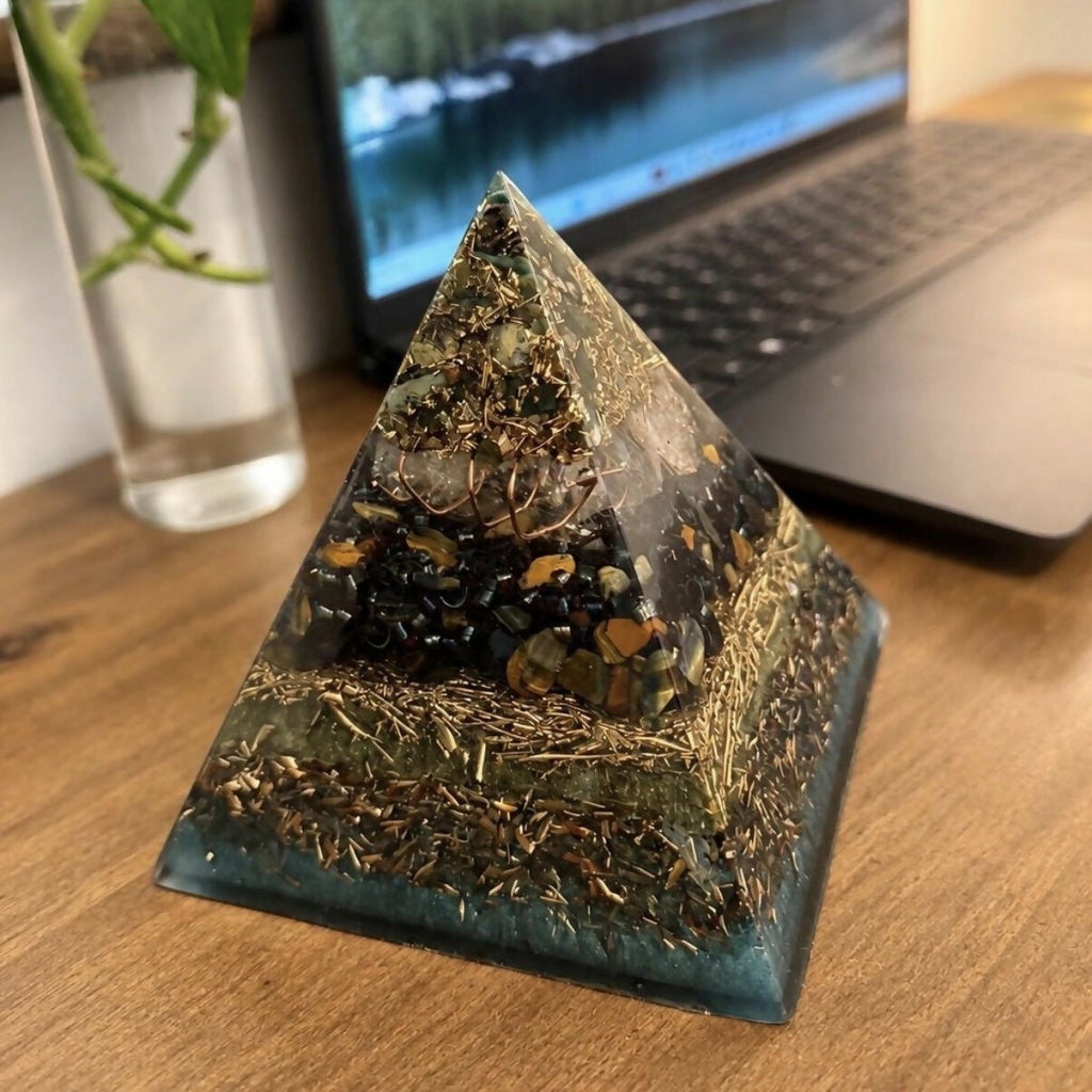 Bloodstone and Kambaba Jasper pyramid by Piezo & Co on a wooden desk beside a laptop, showing clear quartz, dark patterned stones, a band of gold-coloured shavings and a teal base
