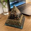 Bloodstone and Kambaba Jasper pyramid by Piezo & Co on a wooden desk beside a laptop, showing clear quartz, dark patterned stones, a band of gold-coloured shavings and a teal base
