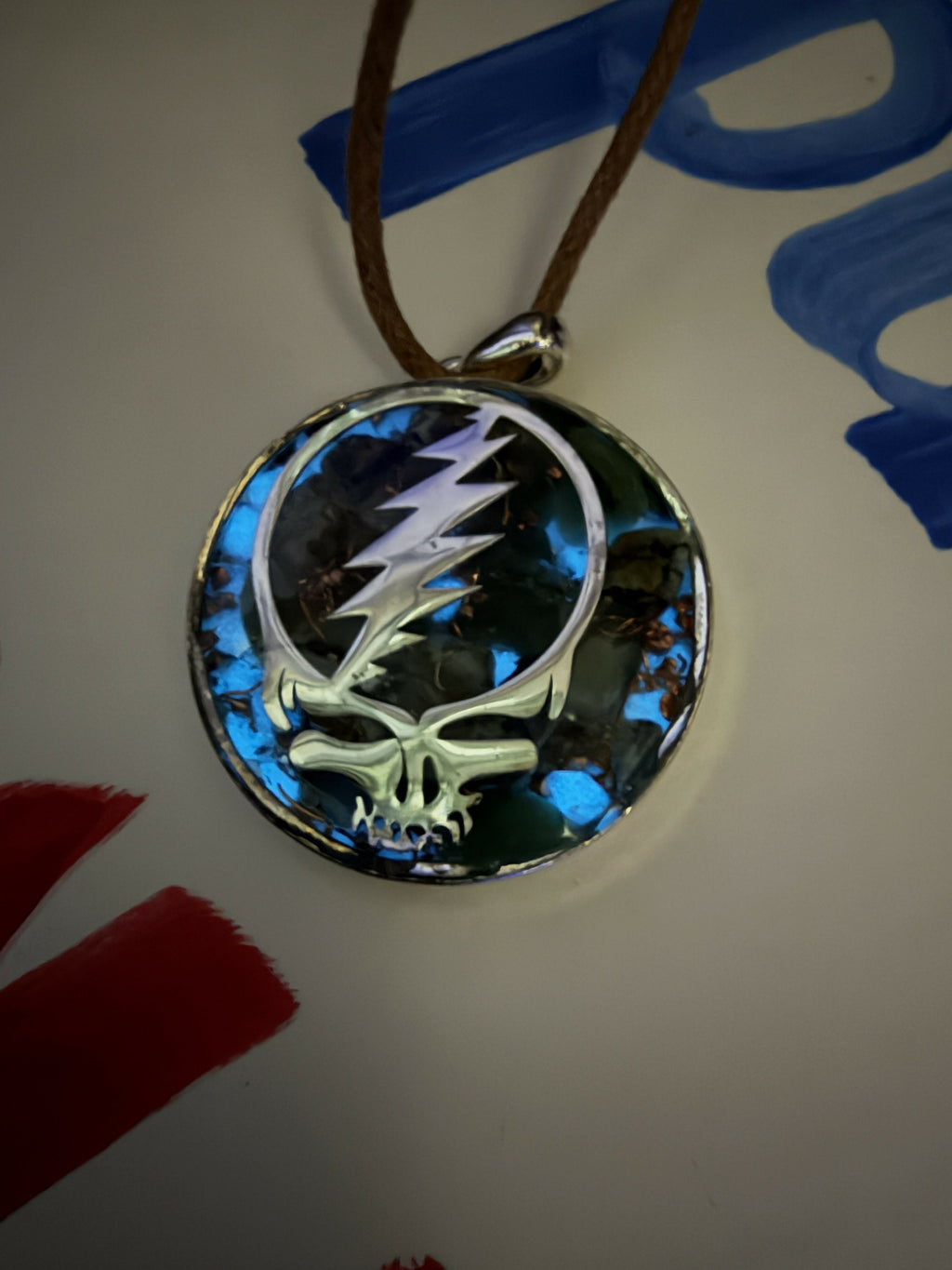 Pendant on a brown cord lying on a white surface, silver skull-and-lightning-bolt design over blue and dark stones