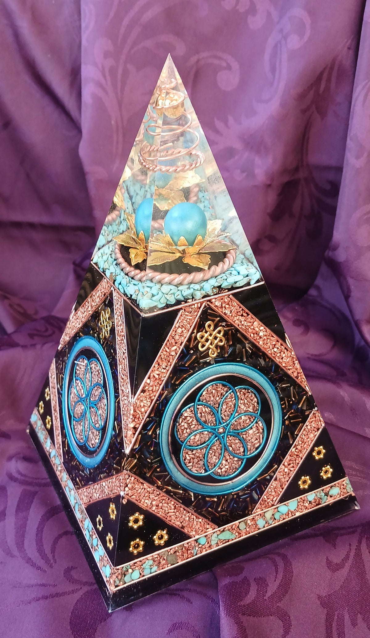 Full-height view of tall turquoise and black orgonite pyramid with Seed of Life decoration on purple fabric.