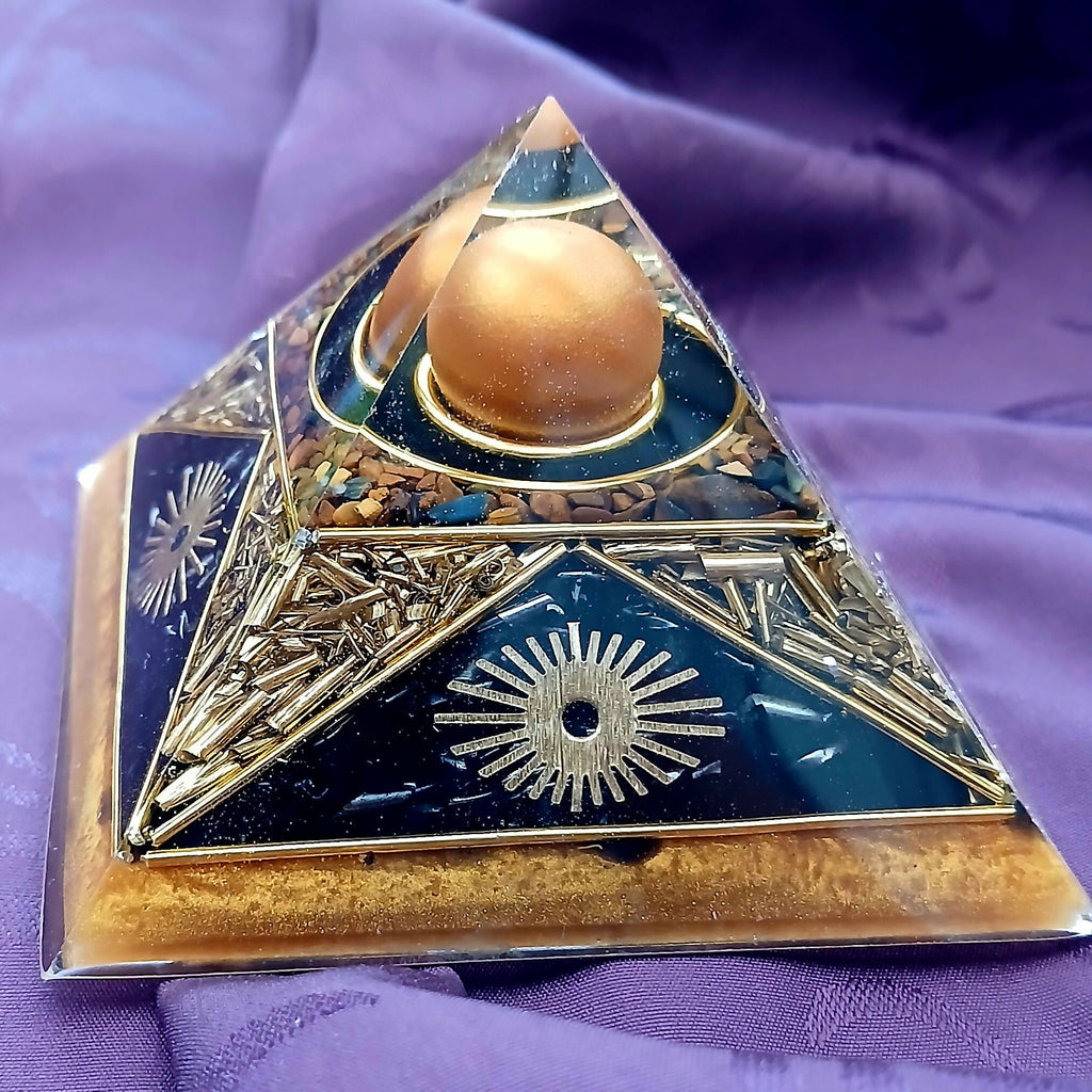 Small Cheops Tiger Eye Pyramid by Orgone Art Yvonne – black and gold pyramid with a large golden-brown sphere at the apex, gold sunburst motifs, golden-brown stone chips and a light tan base