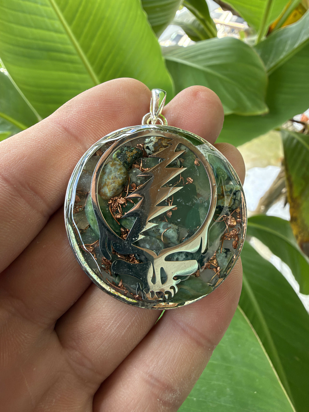 Pendant held in fingers in front of green leaves, showing green stones, copper pieces and a bronze-coloured skull design