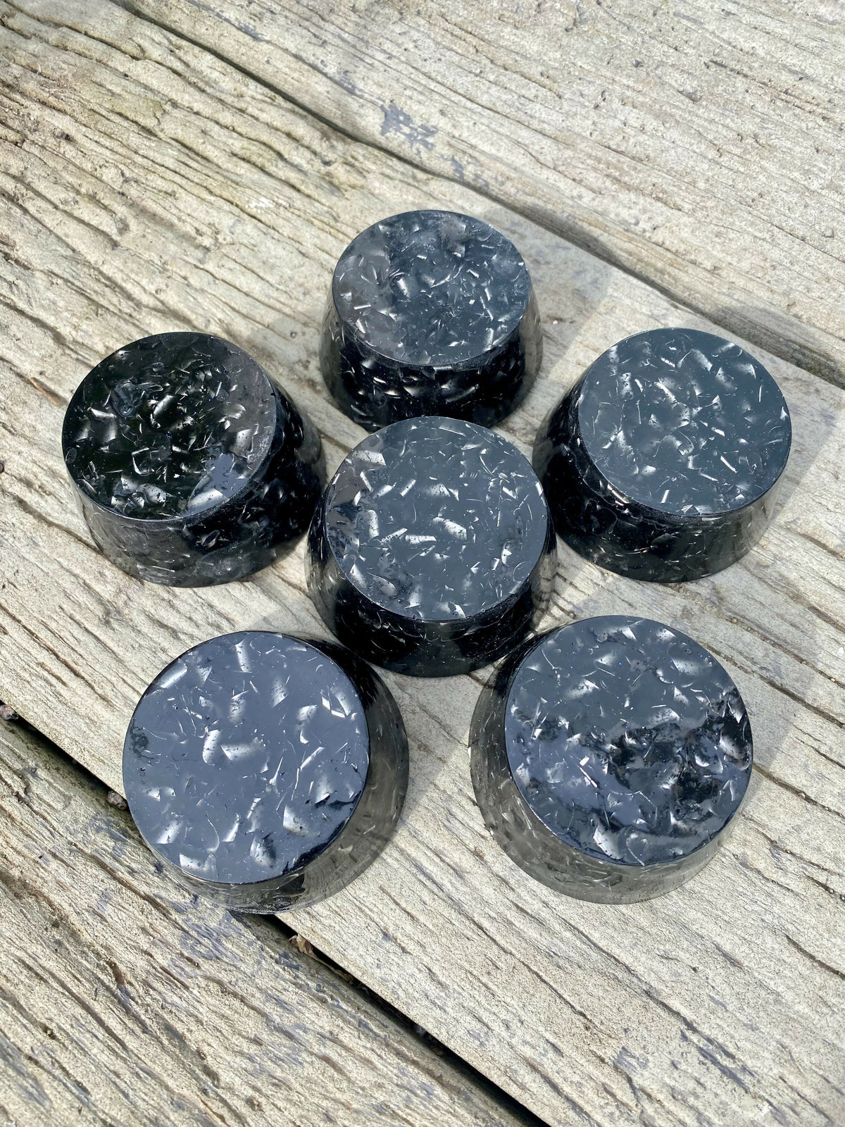 Six round black shungite orgonite towerbusters with silver aluminium flecks, arranged in a circle on a weathered wooden surface