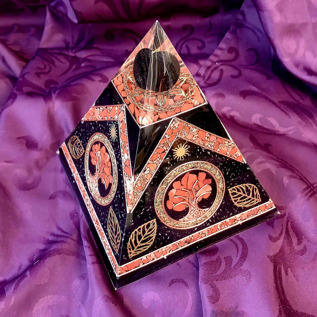 Angled view of Tree of Life orgonite pyramid with red stone chips and metallic leaf decoration on purple fabric.