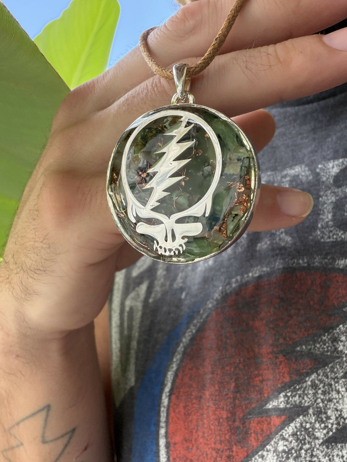 Pendant held up by its cord in front of large green leaves, showing the silver skull-and-lightning-bolt design over green stones