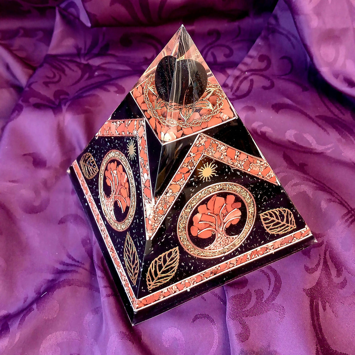 Front angled view of Tree of Life orgonite pyramid with dark panels, red stone chips and clear pointed top.