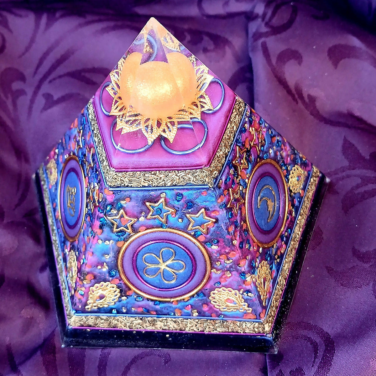 Five-sided blue and purple orgonite pyramid with metallic flower, star and crescent decorations on purple fabric.