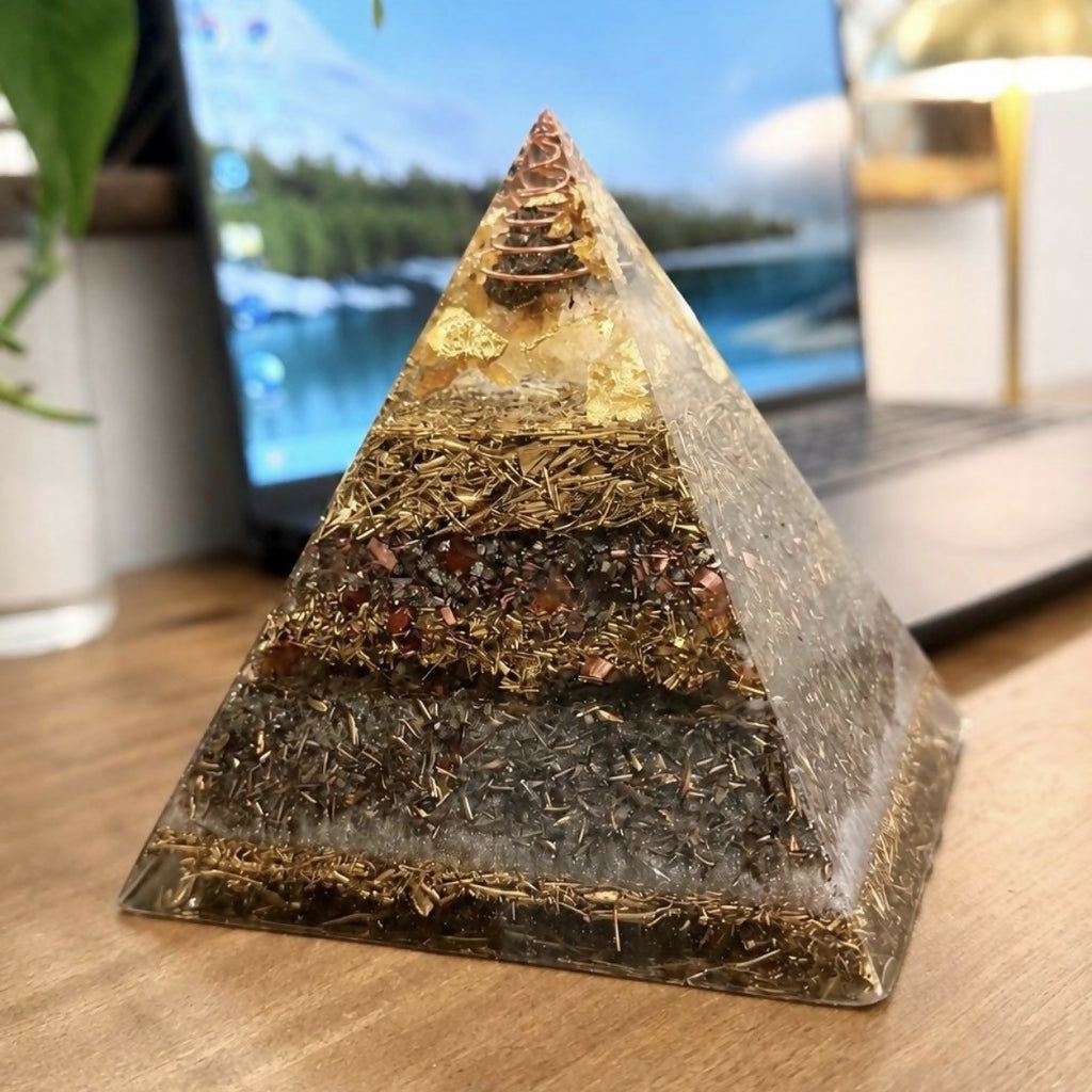 Abundance Orgonite Pyramid by Piezo & Co – layered pyramid with a golden top and copper coil tip on a wooden desk in front of a laptop