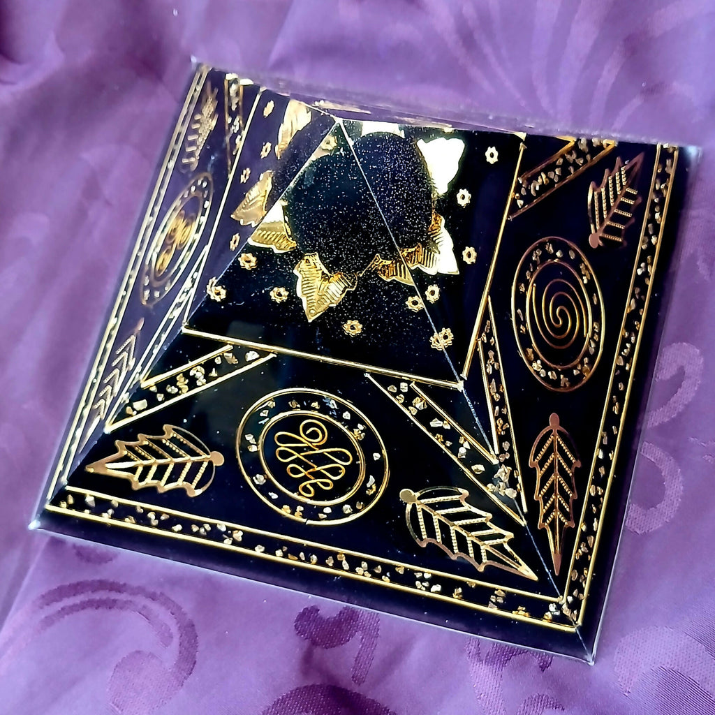 Black Giza Orgonite Pyramid by Orgone Art Yvonne – low black pyramid with gold spirals, gold leaf motifs, gold triangles and a dark sphere at the apex, on purple fabric