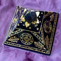 Black Giza Orgonite Pyramid by Orgone Art Yvonne – low black pyramid with gold spirals, gold leaf motifs, gold triangles and a dark sphere at the apex, on purple fabric