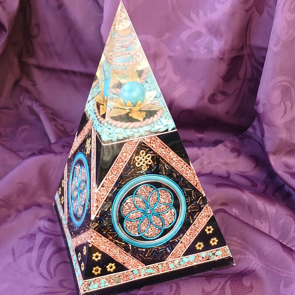 Tall orgonite pyramid with turquoise stone chips, blue Seed of Life panels and copper-coloured borders on purple fabric.