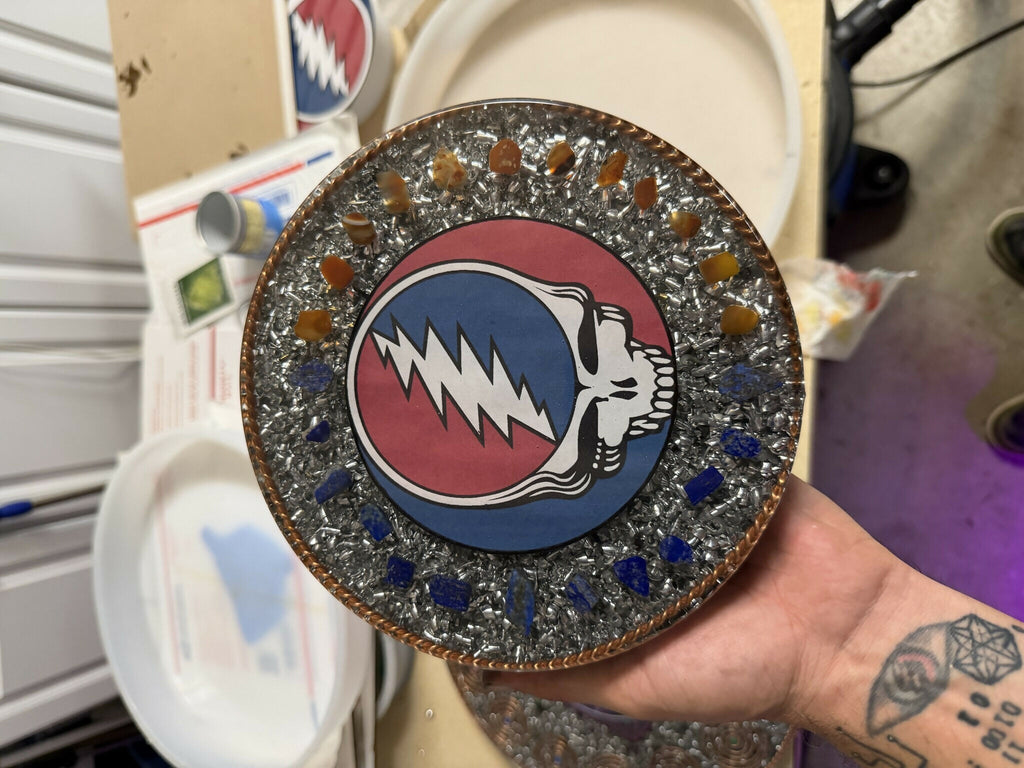 Stealie Lapis & Carnelian Charging Plate by Mark Ethan Designs – round plate with a red, white and blue skull and lightning-bolt design ringed by orange and blue stones and silver pieces, held in a hand