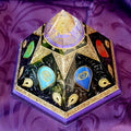 Six-sided orgonite pyramid with colourful chakra symbols, dark panels and gold-coloured decoration on purple fabric.