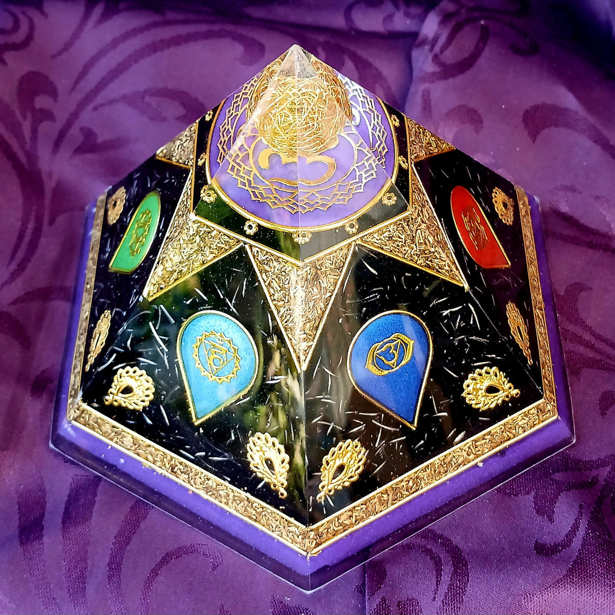 Six-sided orgonite pyramid with colourful chakra symbols, dark panels and gold-coloured decoration on purple fabric.