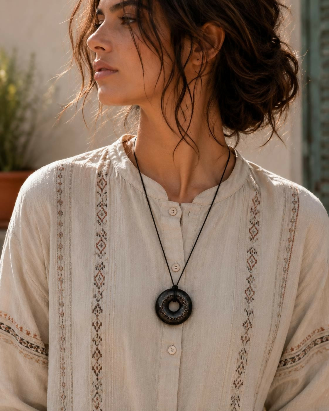The black donut pendant worn on a black cord over a cream embroidered shirt
