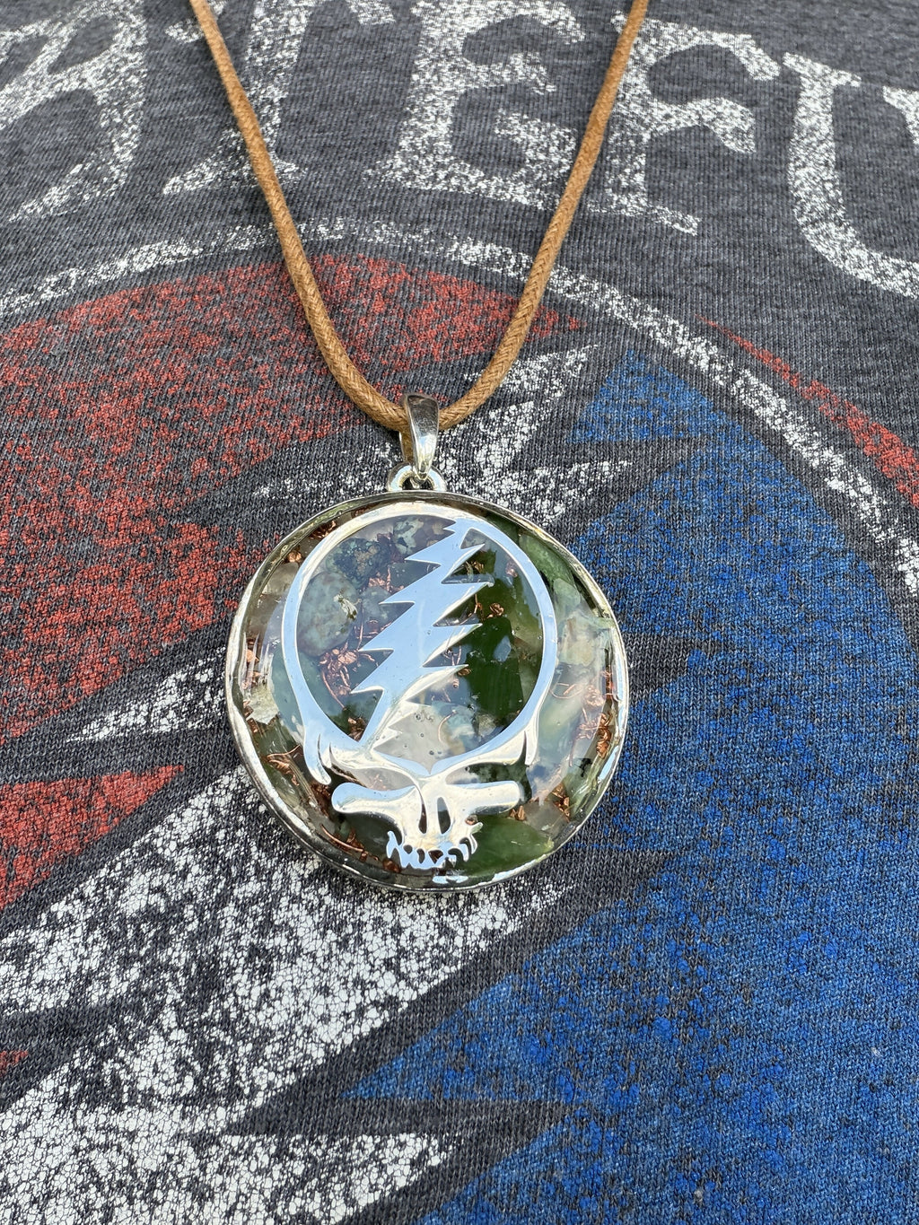 Stealie pendant by Mark Ethan Designs hanging on a tan leather cord, silver skull-and-lightning-bolt cutout over green stones in clear resin