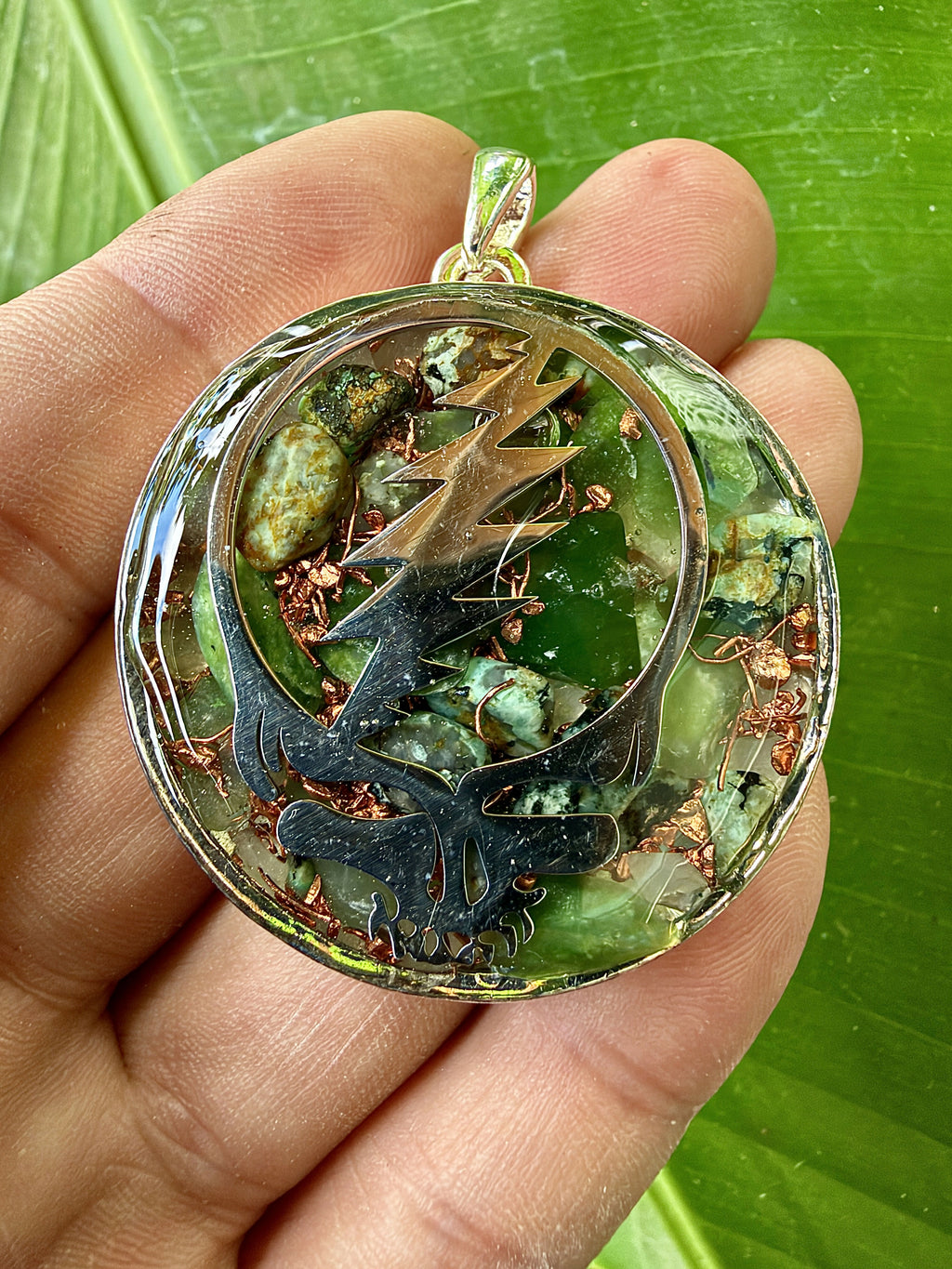 Close view of a pendant held in the palm, bronze-coloured skull-and-lightning-bolt design over green stones and copper pieces