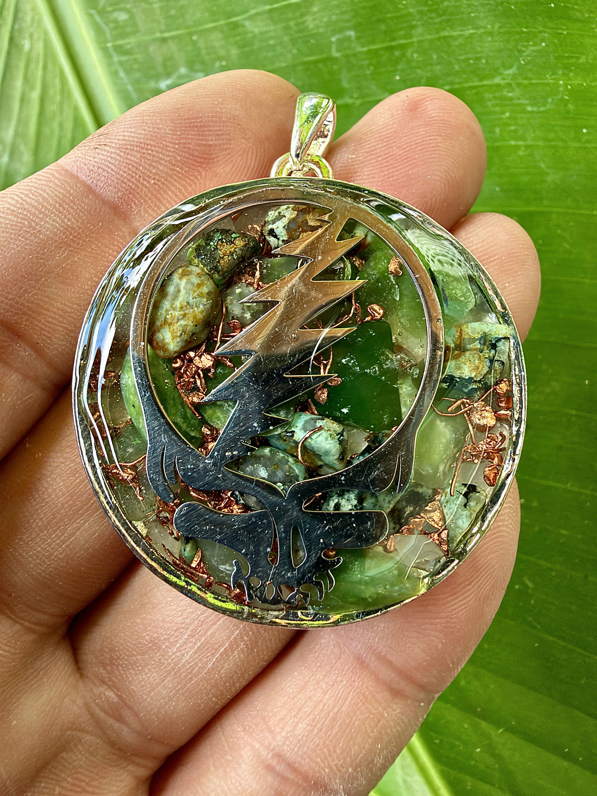 Close view of a pendant held in the palm, bronze-coloured skull-and-lightning-bolt design over green stones and copper pieces