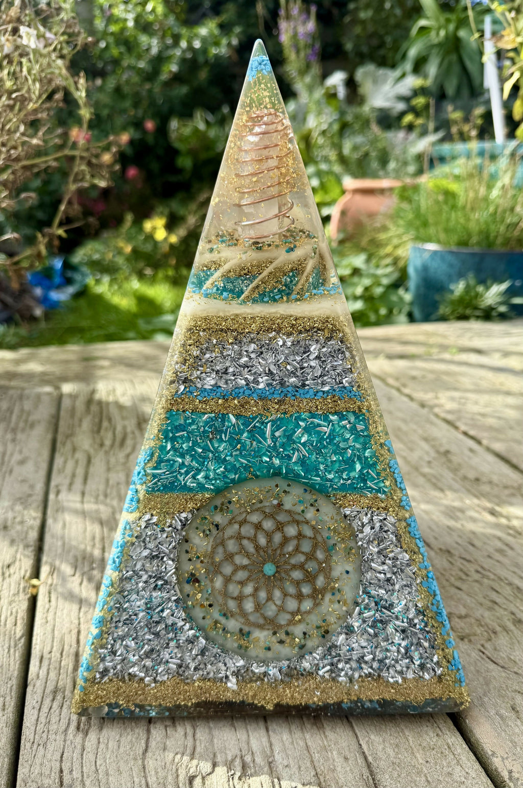 XL Nubian Torus Orgonite Pyramid by Orgone Solutions – tall pyramid with bands of turquoise, silver and gold glitter, a gold Flower of Life disc and a clear point at the top, outdoors on wood