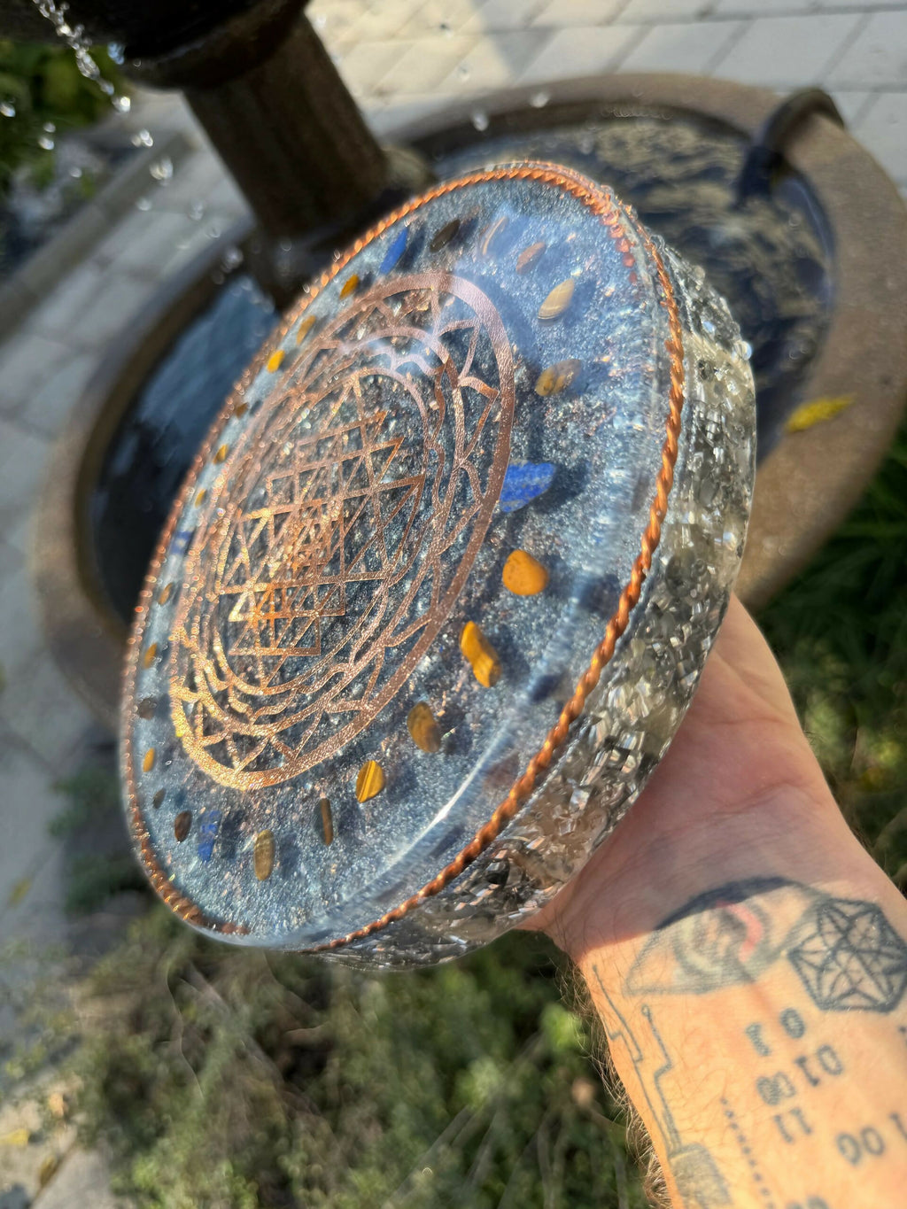 Angled top view of the plate held outdoors, showing the copper Sri Yantra design and a ring of blue and orange stones